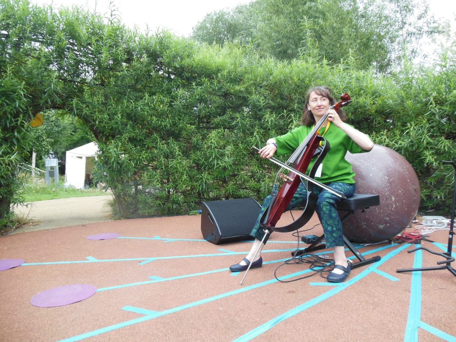 PS Arch Joanne sits in a public garden playing electric cello, with conductive tape on the floor around her