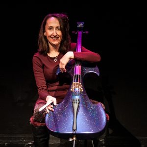 Joanne smiles with one arm hooked around her sparkly purple cello