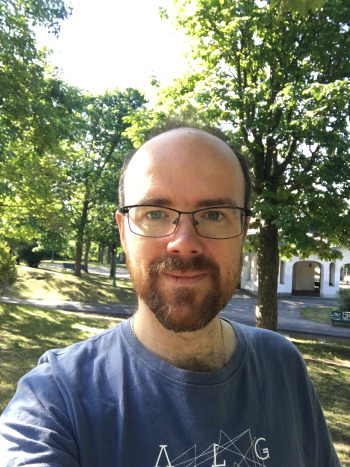 White man with glasses and a dark brown beard looks into the camera wearing a blue t-shirt with trees in the background
