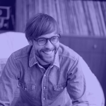 A bearded white man wearing glasses and a denim shirt sits, smiling and looking away from the camera