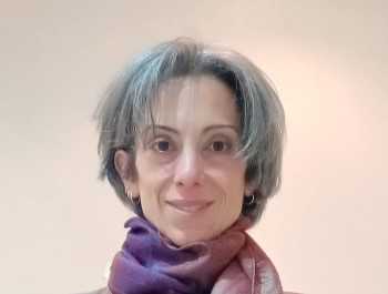 A coloured profile photo of a white female, with short black and white hair, wearing a bright purple-beige coloured scarf around the neck and smiling to the camera.
