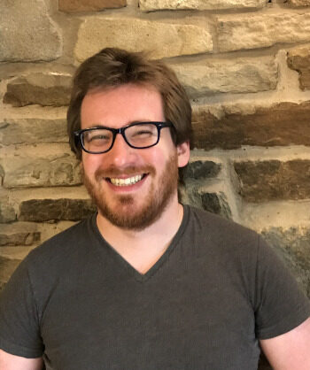 A person with short brown hair and beard, and blue glasses, wearing a grey t-shirt stands in front of a brick wall, smiling