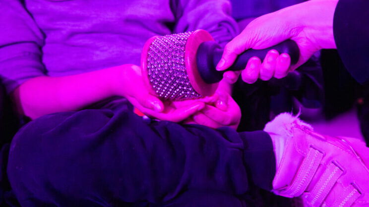 A student cups her hands on her lap to feel the texture of a cabasa, a percussive instrument, being offered to the student by a member of staff. The cabasa consists of loops of steel ball chain wrapped around a wooden cylinder, fixed to a black plastic handle. The photo appears tinted as the room is illuminated by a pinky-purple light.