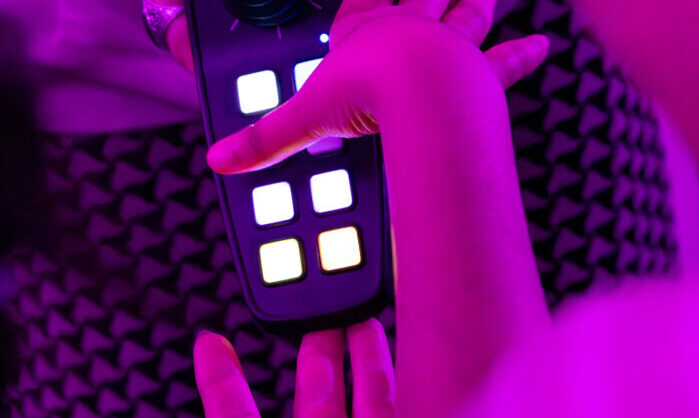 A student’s hand is pressing a button on a MIDI instrument called CMPSR. The CMPSR is a black handheld device with two rows of four buttons and a joystick above, akin to a wheelchair controller. The CMPSR is being held up for the student to interact with by Valeria, a member of the creative team, and a teacher. The photo appears tinted as the room is illuminated by a pinky-purple light.