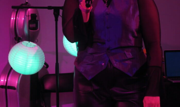A photo of Valeria singing at Rich Mix. Valeria is a white person with blonde hair. Their eyes are closed, they look like they're shouting. Valeria is wearing a black mesh long sleeve top, an iridescent waist coat and black pleather trousers. The lighting is purple.