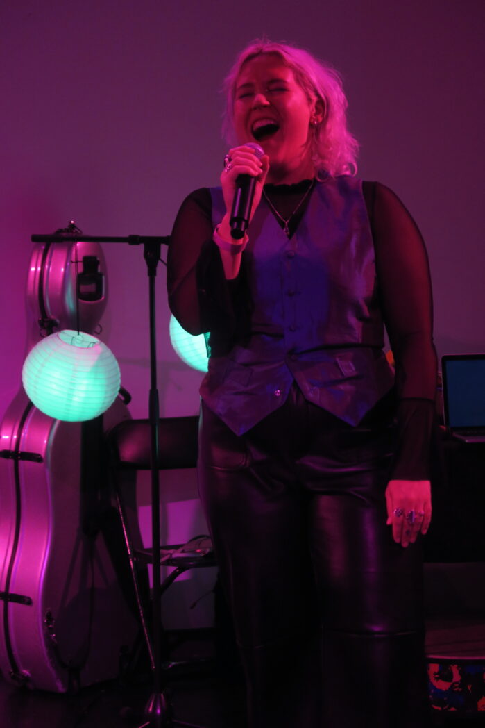 A photo of Valeria singing at Rich Mix. Valeria is a white person with blonde hair. Their eyes are closed, they look like they're shouting. Valeria is wearing a black mesh long sleeve top, an iridescent waist coat and black pleather trousers. The lighting is purple.