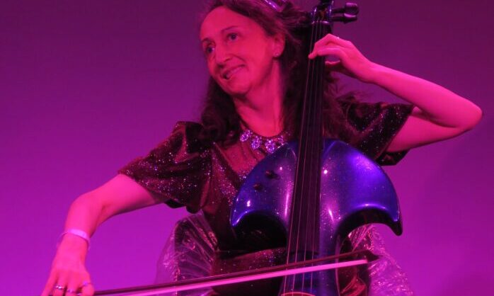 A photo of Jo-anne playing cello at Rich Mix. Jo-anne is a white woman in her 50's seated with her purple sparkling cello. Her right arm is extended, drawing the bow across a string. Her left arm is held up and fingers are on the strings. She is wearing a burgundy short sleeved sparkly dress, a silver sparkly necklace and long brown hair. She is looking to one side smiling. The background lighting is purple.
