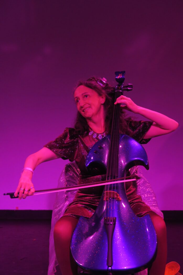 A photo of Jo-anne playing cello at Rich Mix. Jo-anne is a white woman in her 50's seated with her purple sparkling cello. Her right arm is extended, drawing the bow across a string. Her left arm is held up and fingers are on the strings. She is wearing a burgundy short sleeved sparkly dress, a silver sparkly necklace and long brown hair. She is looking to one side smiling. The background lighting is purple.