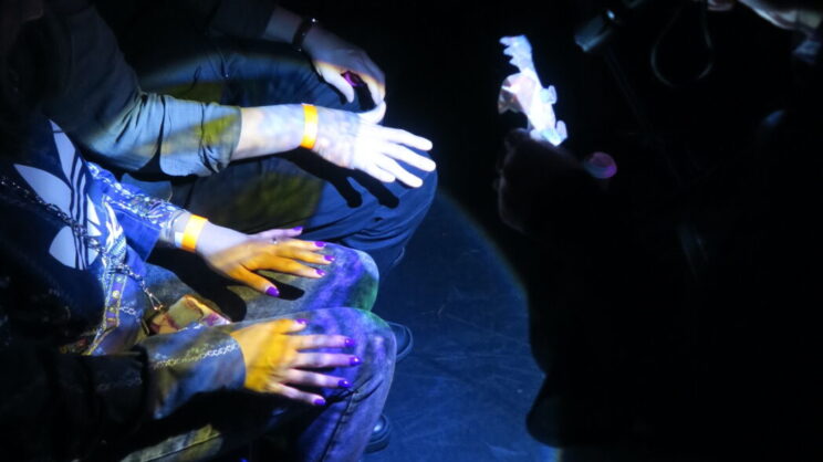 A photo of a shadow dragon puppet appearing on the hands of several audience members. A torch shines light through the puppet, casting yellow, purple and green light on their hands.