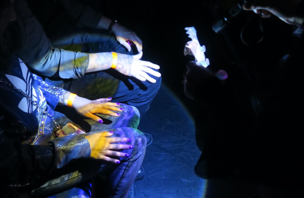 A photo of a shadow dragon puppet appearing on the hands of several audience members. A torch shines light through the puppet, casting yellow, purple and green light on their hands.