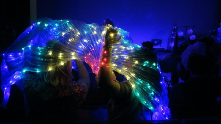 A photo of LED light up wings being held up by audience members. The lights are red, yellow, green and blue on translucent fabric.