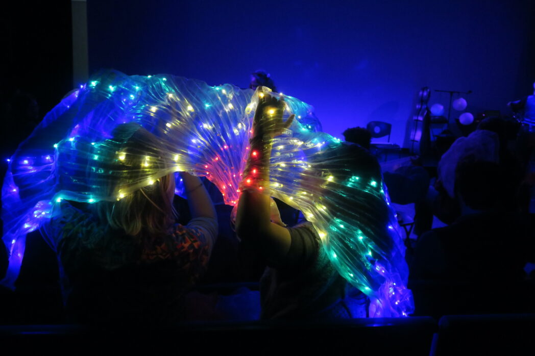 A photo of LED light up wings being held up by audience members. The lights are red, yellow, green and blue on translucent fabric.